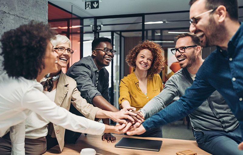 Equipe de seis adultos de varias etnias, idades e generos juntando as mãos comemorativamente ao redor de uma mesa de reunião em um ambiente corporativo despojado. Luz tênue vindo do topo. Todos sorriem e se olham nos olhos com respeito e dignidade.